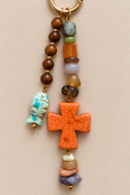 A beaded keychain charm featuring a large orange crackled cross, a turquoise fish-shaped bead, brown wooden beads, and various colorful stone beads on a gold-tone ring, set against a solid beige background.