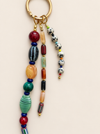 A bohemian-style beaded keychain with a gold-tone split ring, featuring multiple strands of colorful glass beads in red, green, orange, black, white, and blue on a light beige background.
