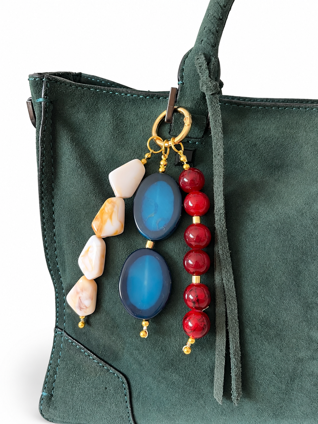 A close-up of a dark green suede handbag featuring a decorative keychain or bag charm with blue agate stones, red beads, and natural stone pieces on gold-toned rings.