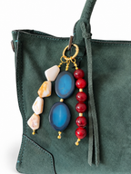 A close-up of a dark green suede handbag featuring a decorative keychain or bag charm with blue agate stones, red beads, and natural stone pieces on gold-toned rings.