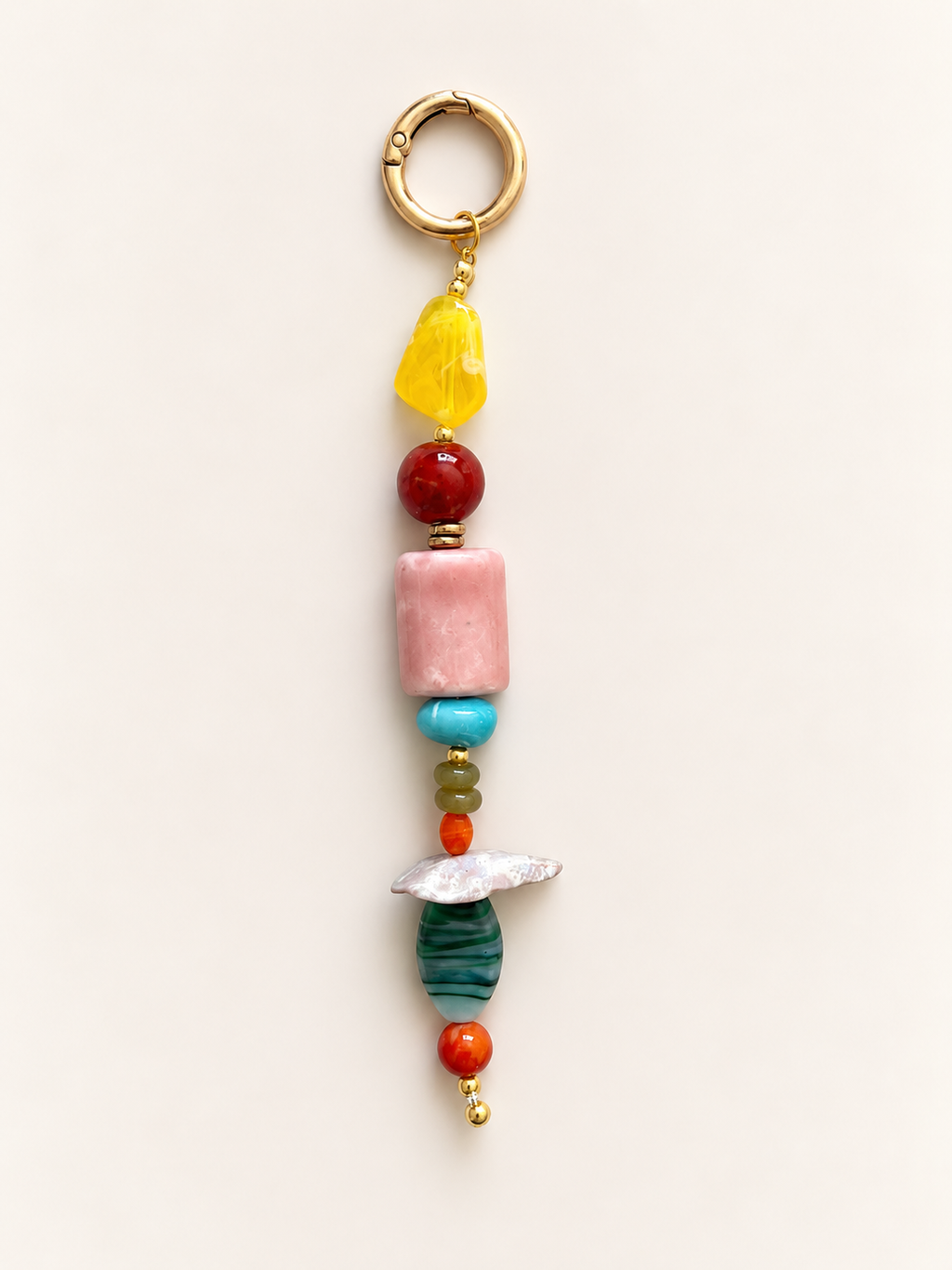 A handmade beaded keychain charm featuring a gold-tone split ring, a yellow translucent stone, a red bead, a pink rectangular stone, a turquoise bead, smaller green and orange beads, a white shell, a dark green striped stone, and a small gold bead, displayed on a light beige background.