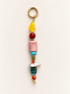 A handmade beaded keychain charm featuring a gold-tone split ring, a yellow translucent stone, a red bead, a pink rectangular stone, a turquoise bead, smaller green and orange beads, a white shell, a dark green striped stone, and a small gold bead, displayed on a light beige background.
