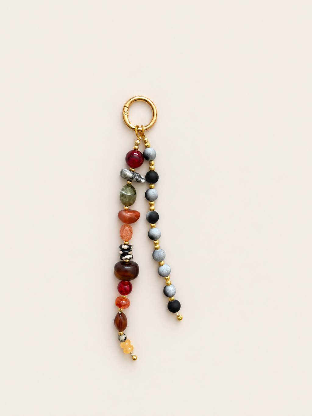 A beaded tassel charm with a gold-tone ring and split strands of polished gemstone beads in red, brown, gray, black, orange, and yellow, shown on a light beige background.