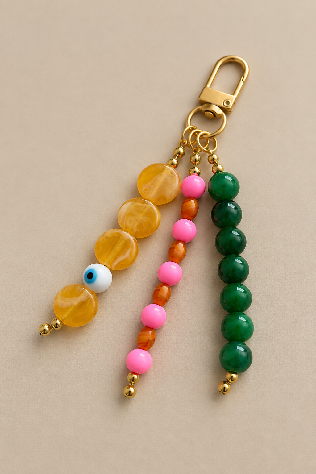 A gold-tone keychain bag charm featuring three beaded strands: one with translucent yellow beads and a white evil eye bead, one with pink and orange beads, and one with dark green beads, all displayed on a neutral beige background.