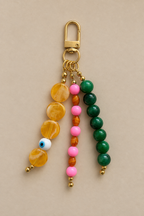 A gold-tone keychain bag charm featuring three hanging beaded strands: one with yellow marbled beads and an evil eye bead, one with pink and orange beads, and one with green beads, all on a beige background.