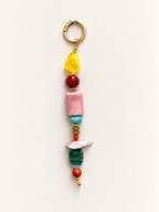 A beaded keychain charm featuring a gold split ring, a translucent yellow stone, a red bead, a pink rectangular stone, turquoise beads, a white shell, and green and orange beads on a plain off-white background.