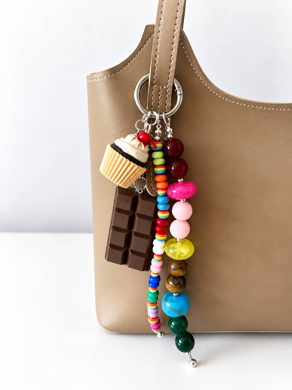 A tan leather shoulder bag with a colorful beaded keychain charm featuring a cupcake, chocolate bar, and round beads, shown against a white background.