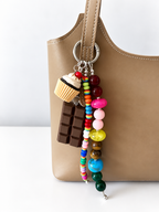 A tan leather shoulder bag with a colorful beaded keychain charm featuring a cupcake, chocolate bar, and round beads, shown against a white background.