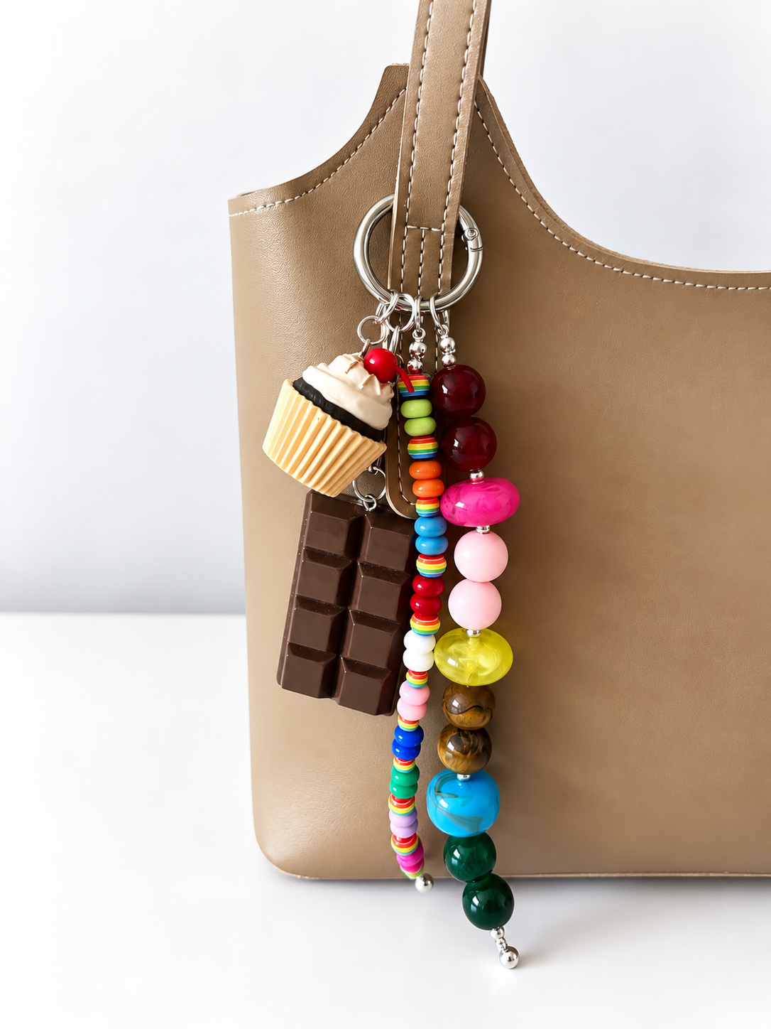 A tan leather shoulder bag with a colorful beaded keychain charm featuring a cupcake, chocolate bar, and round beads, shown against a white background.