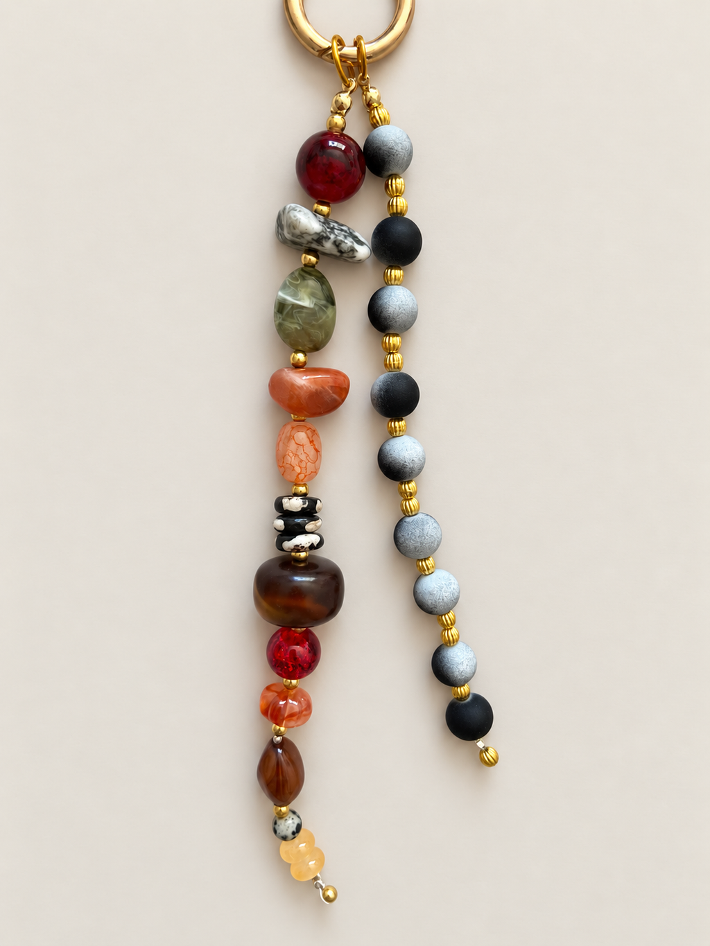 A beaded keychain with a gold-tone split ring, featuring two hanging strands of polished gemstone beads in red, green, orange, brown, black, gray, and white, separated by small gold spacer beads, on a light beige background.