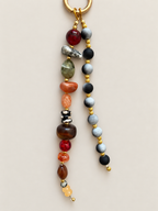 A beaded keychain with a gold-tone split ring, featuring two hanging strands of polished gemstone beads in red, green, orange, brown, black, gray, and white, separated by small gold spacer beads, on a light beige background.