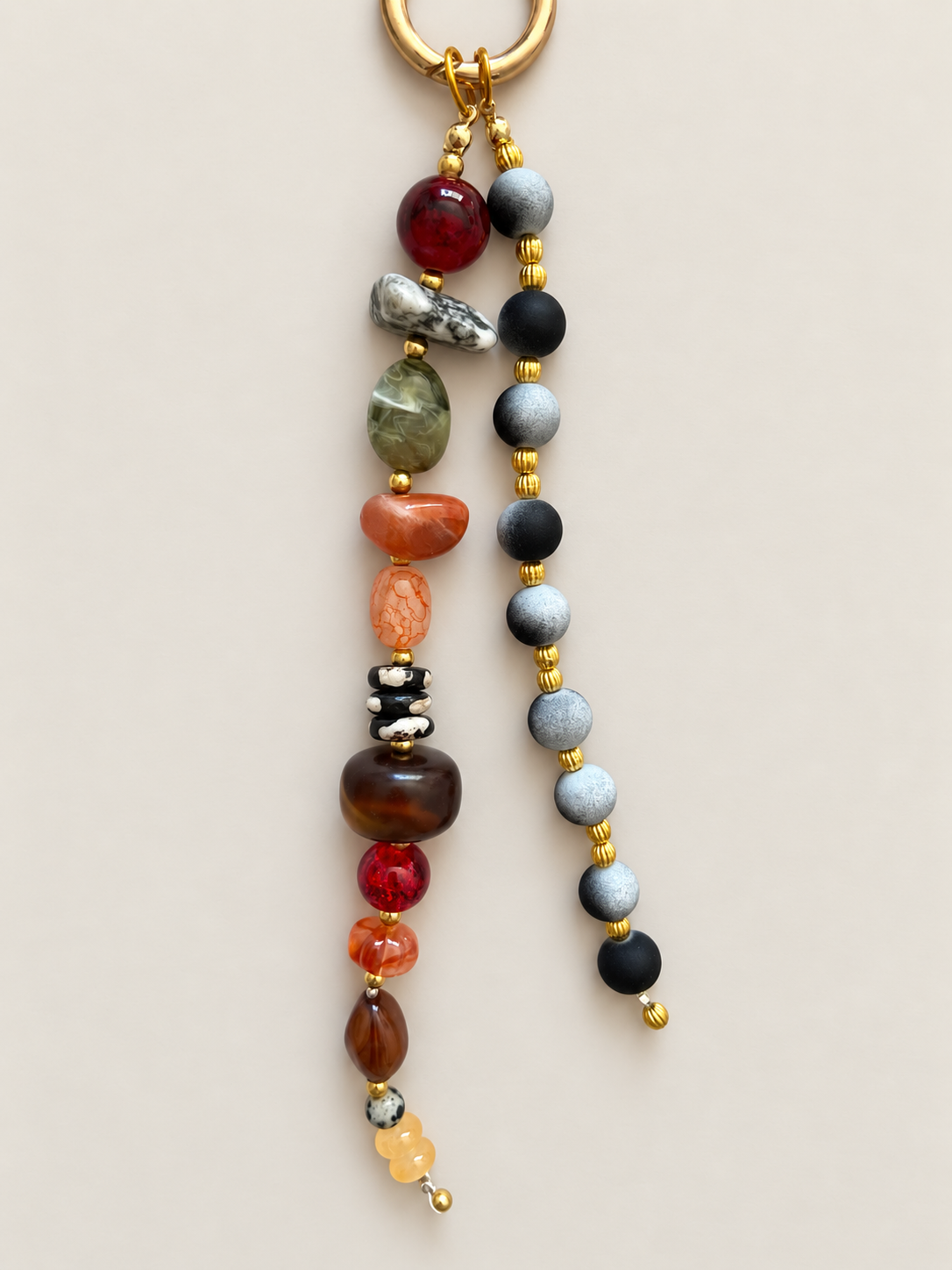A beaded keychain with a gold-tone split ring, featuring two hanging strands of polished gemstone beads in red, green, orange, brown, black, gray, and white, separated by small gold spacer beads, on a light beige background.