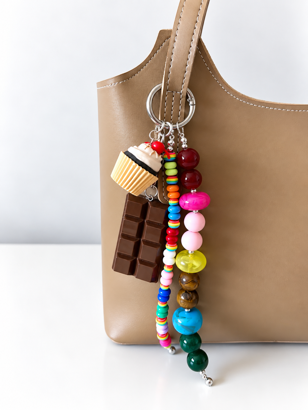 A close-up of a tan leather tote bag with a colorful beaded keychain charm hanging from the handle. The charm includes a chocolate bar, a cupcake, and multiple strands of colorful beads.