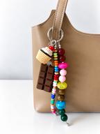 A close-up of a tan leather tote bag with a colorful beaded keychain charm hanging from the handle. The charm includes a chocolate bar, a cupcake, and multiple strands of colorful beads.