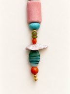 A vertical strand of colorful artisanal beads on a plain off-white background. The set features a large pink cylindrical bead, a turquoise disc, small olive green and orange beads, a textured white shell, a green and blue striped teardrop bead, another orange bead, and small gold spacer beads at the bottom.