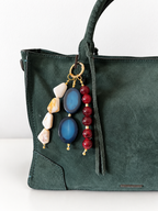 A dark green suede tote bag with a gold-tone ring clasp, adorned with a bohemian-style keychain featuring blue agate stones, cream-colored beads, and red beads.