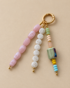 A beaded keychain charm with a gold split ring, featuring three strands of polished beads: light pink, white, and a multicolor pastel mix with a large marbled green-blue bead, yellow spacer beads, and light blue beads, all on a warm beige background.