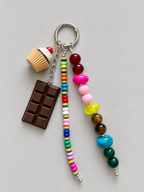 A beaded keychain with three dangling strands: a chocolate bar charm, a cupcake charm, and two colorful bead strands. The keychain is displayed on a neutral gray background.