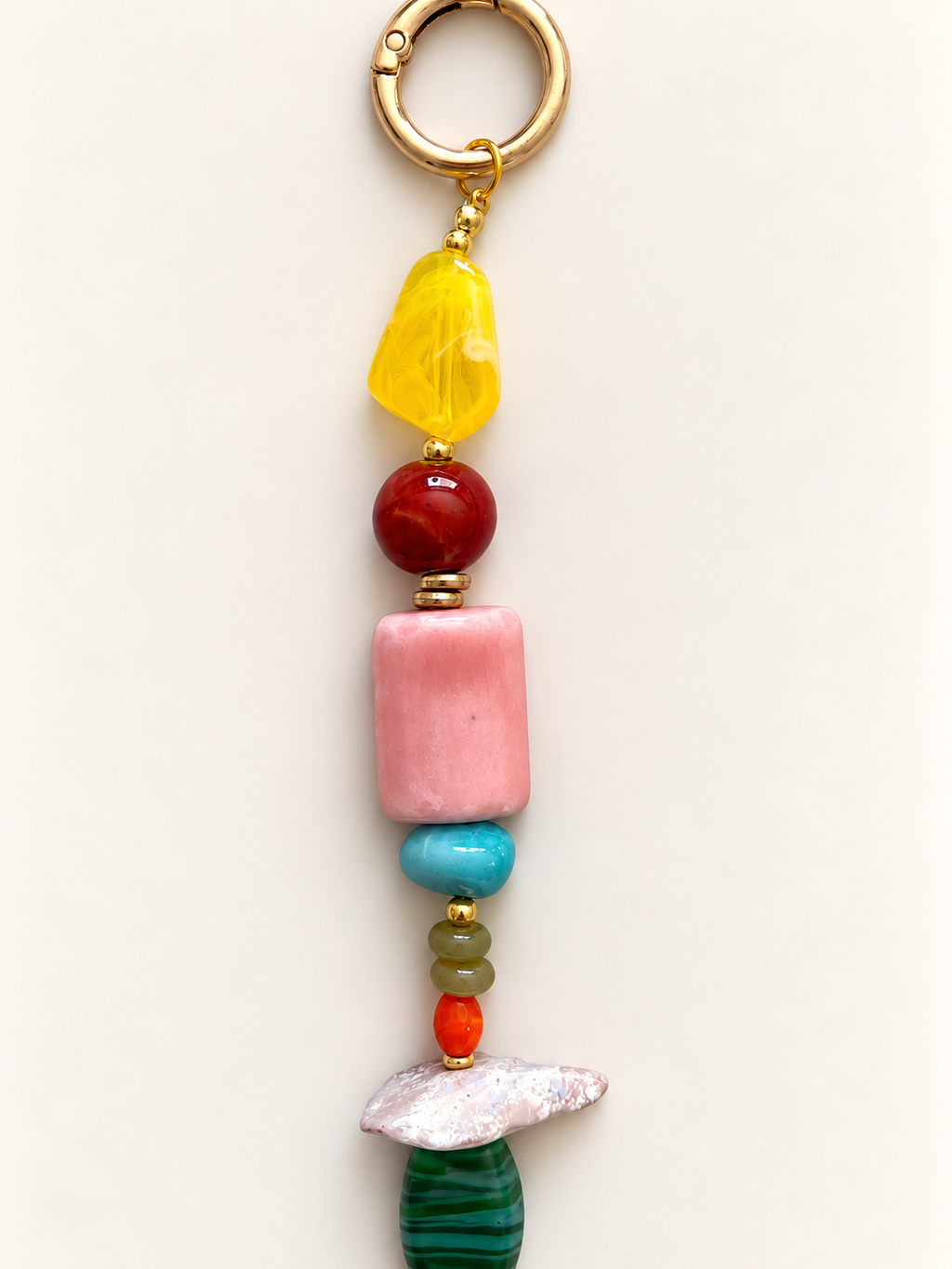 A beaded keychain charm featuring a gold split ring, a faceted yellow stone, a red bead, a pink cylindrical bead, a turquoise bead, small green and orange beads, a white textured stone, and a green striped stone, displayed on a clean off-white background.