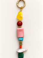 A beaded keychain charm featuring a gold split ring, a faceted yellow stone, a red bead, a pink cylindrical bead, a turquoise bead, small green and orange beads, a white textured stone, and a green striped stone, displayed on a clean off-white background.