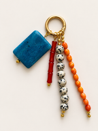 A beaded keychain with a gold-tone split ring, featuring a rectangular blue stone charm, a red cylindrical bead, white and black speckled beads, and orange beads on a light beige background.