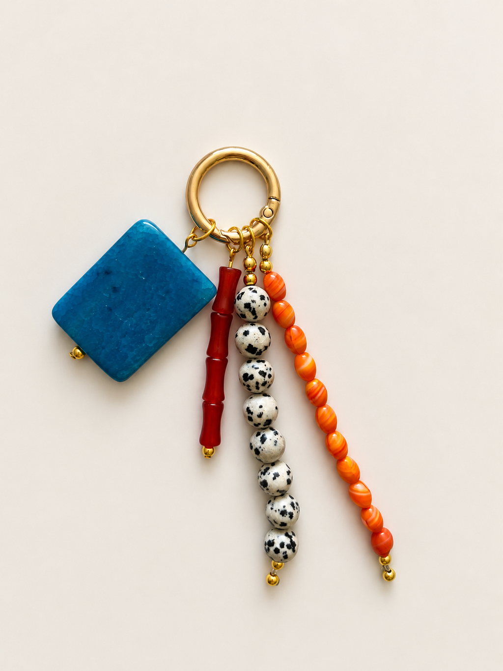 A bohemian-style keychain featuring a square blue stone charm, a red cylindrical bead, white and black spotted beads, and orange round beads, all attached to a gold-tone split ring on a light beige background.
