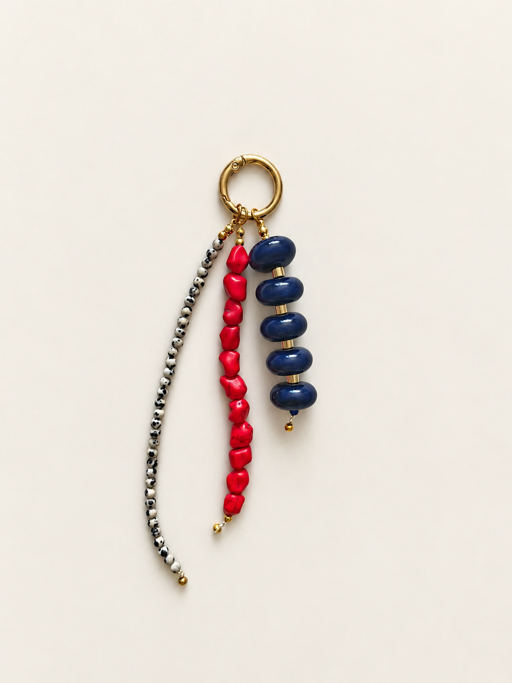 A beaded keychain featuring a gold-tone split ring, a strand of dark blue glossy beads, a strand of bright red heart-shaped beads, and a long strand of small metallic silver beads, all displayed on a light cream background.