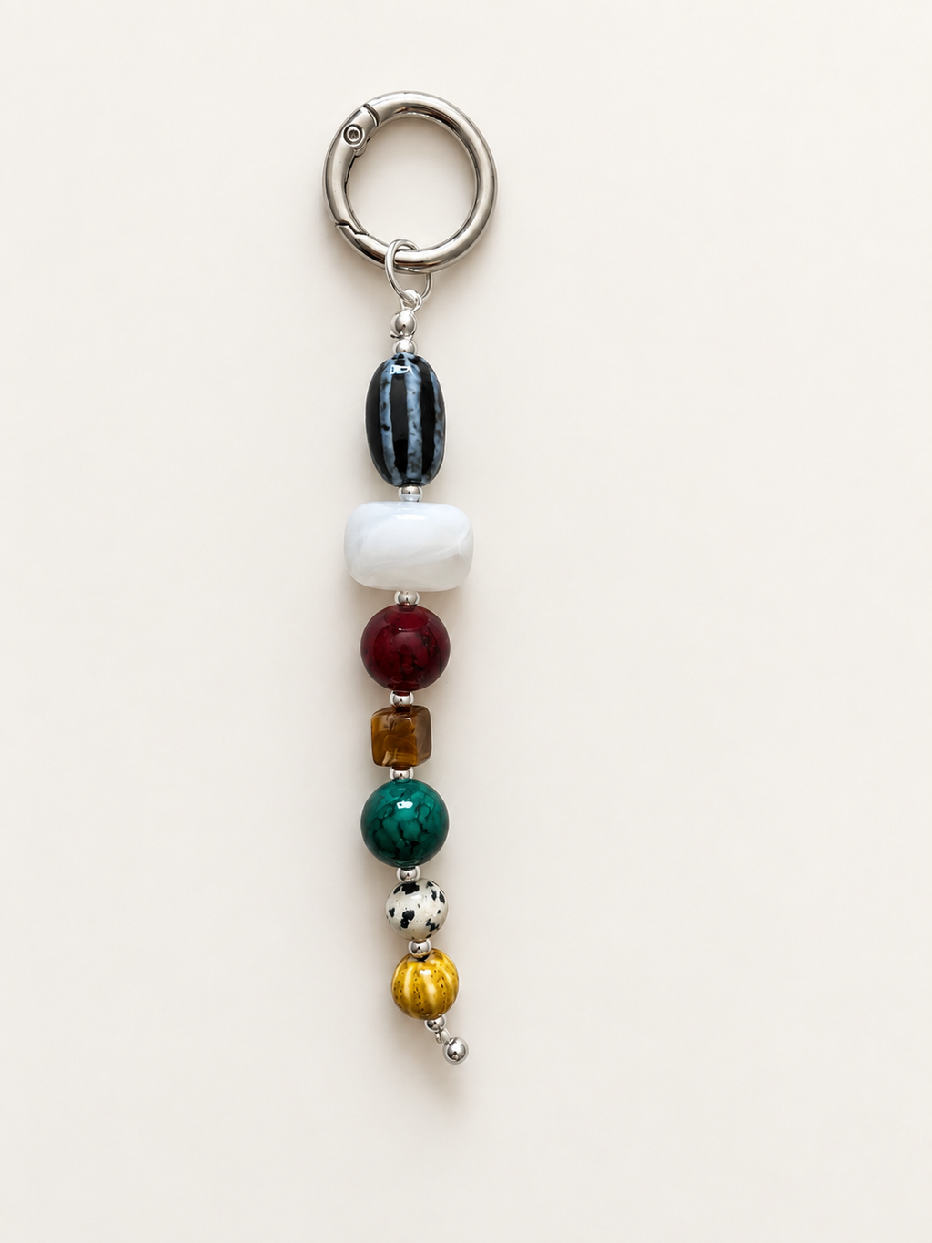 A beaded keychain charm featuring a silver metal split ring and a vertical line of polished beads in black, white, burgundy, amber, green, silver, and gold, displayed on a light cream background.