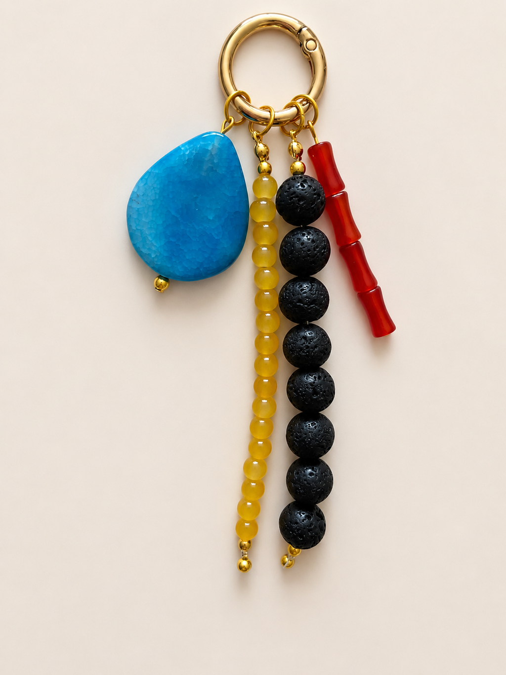 A beaded keychain charm with a gold-tone split ring, featuring a turquoise blue stone, yellow beads, black lava beads, and a red beaded segment on a light beige background.