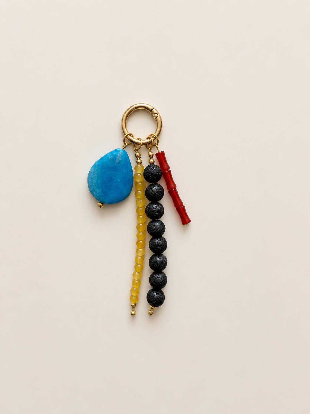 A handmade beaded keychain featuring a gold-tone split ring with three dangling charms: a textured blue stone, a strand of black beads, a strand of yellow beads, and a small red bamboo charm, all on a light beige background.