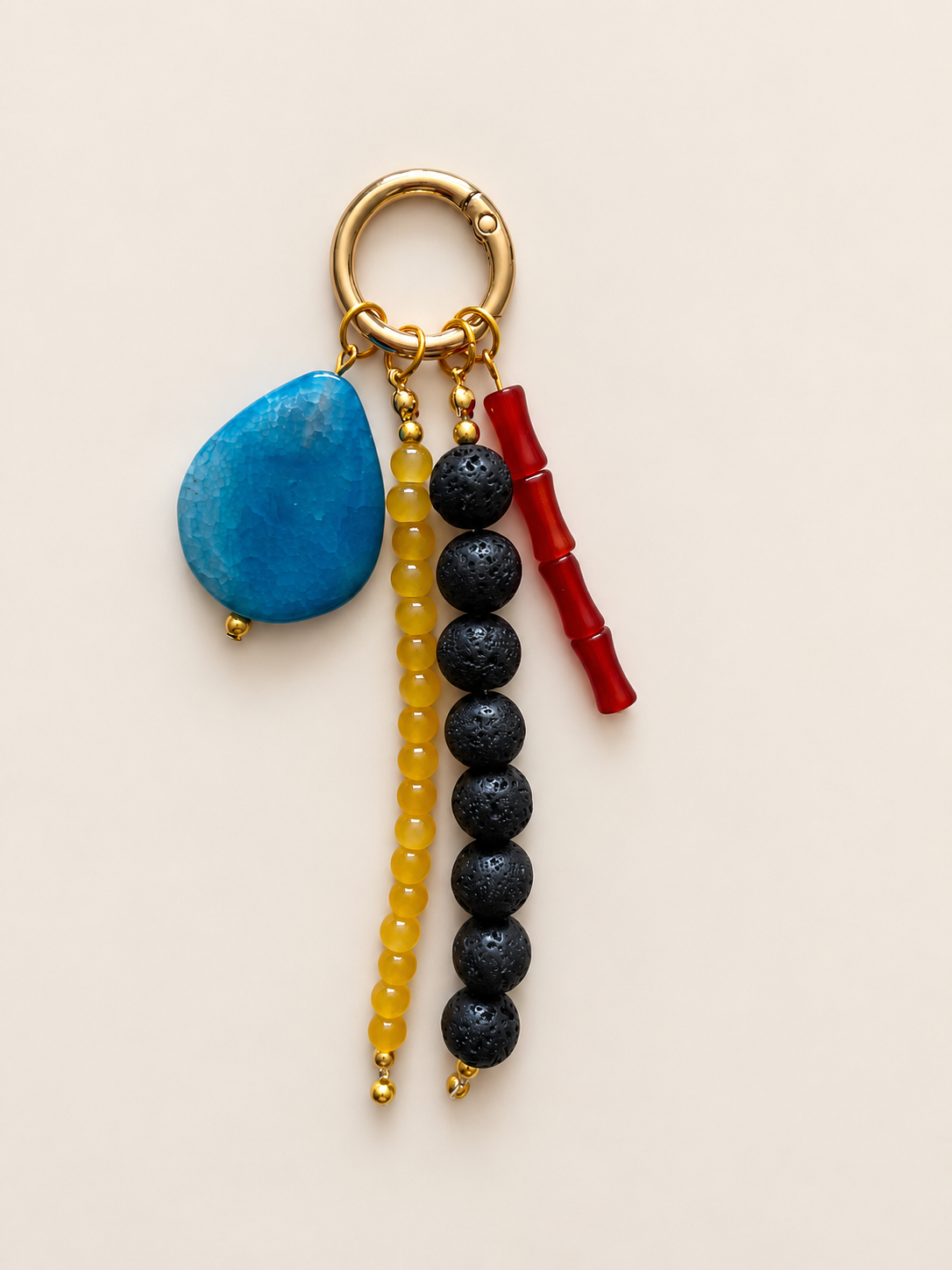 A beaded keychain with a gold-tone split ring, featuring a turquoise blue stone, a strand of yellow beads, a strand of black lava beads, and a red bamboo charm, set against a solid light beige background.