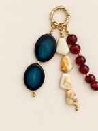 A gold-tone keychain charm featuring two dark blue oval stone pendants, three cream and marbled stone pieces, and a strand of deep red beads, displayed on a light beige background.