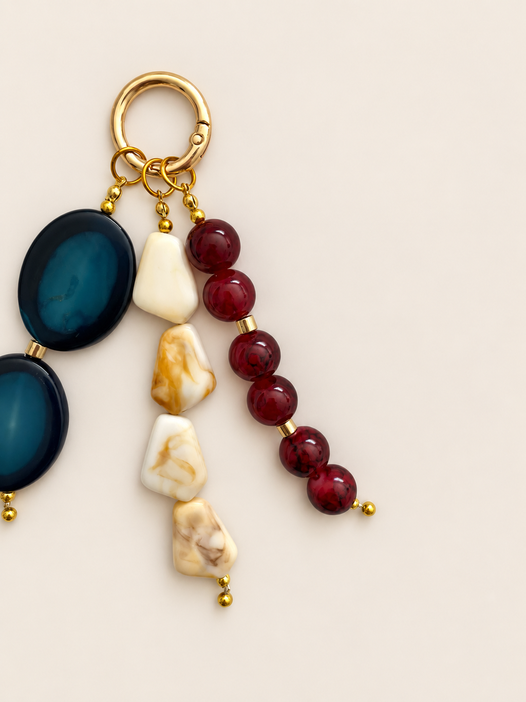 A beaded keychain charm with a gold-tone split ring, featuring dark blue oval beads, cream and marbled stone pieces, and dark red round beads separated by small gold spacers, on a light beige background.