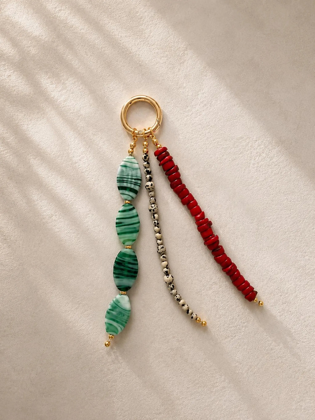 A beaded keychain or bag charm with a gold-tone ring, featuring three dangling strands: green marbled beads, a silver-and-black bead strand, and a red beaded strand, displayed on a textured cream fabric with soft shadows.
