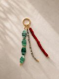 A beaded keychain or bag charm with a gold-tone ring, featuring three dangling strands: green marbled beads, a silver-and-black bead strand, and a red beaded strand, displayed on a textured cream fabric with soft shadows.