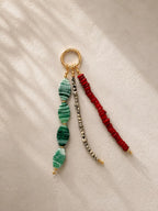 A beaded keychain or bag charm with a gold-tone ring, featuring three dangling strands: green marbled beads, a silver-and-black bead strand, and a red beaded strand, displayed on a textured cream fabric with soft shadows.