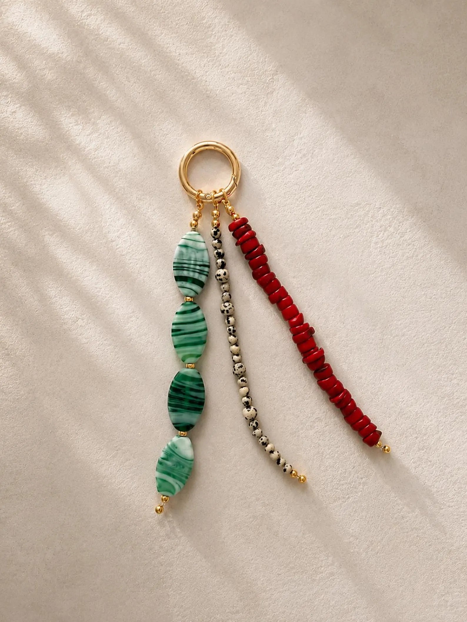 A beaded keychain or bag charm with a gold-tone ring, featuring three dangling strands: green marbled beads, a silver-and-black bead strand, and a red beaded strand, displayed on a textured cream fabric with soft shadows.